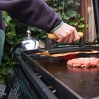 Primer plano de una prensa para carne con mango de madera utilizándose sobre la plancha del asador Napoleon Freestyle 24 - Ferretería La Palma - 1890000