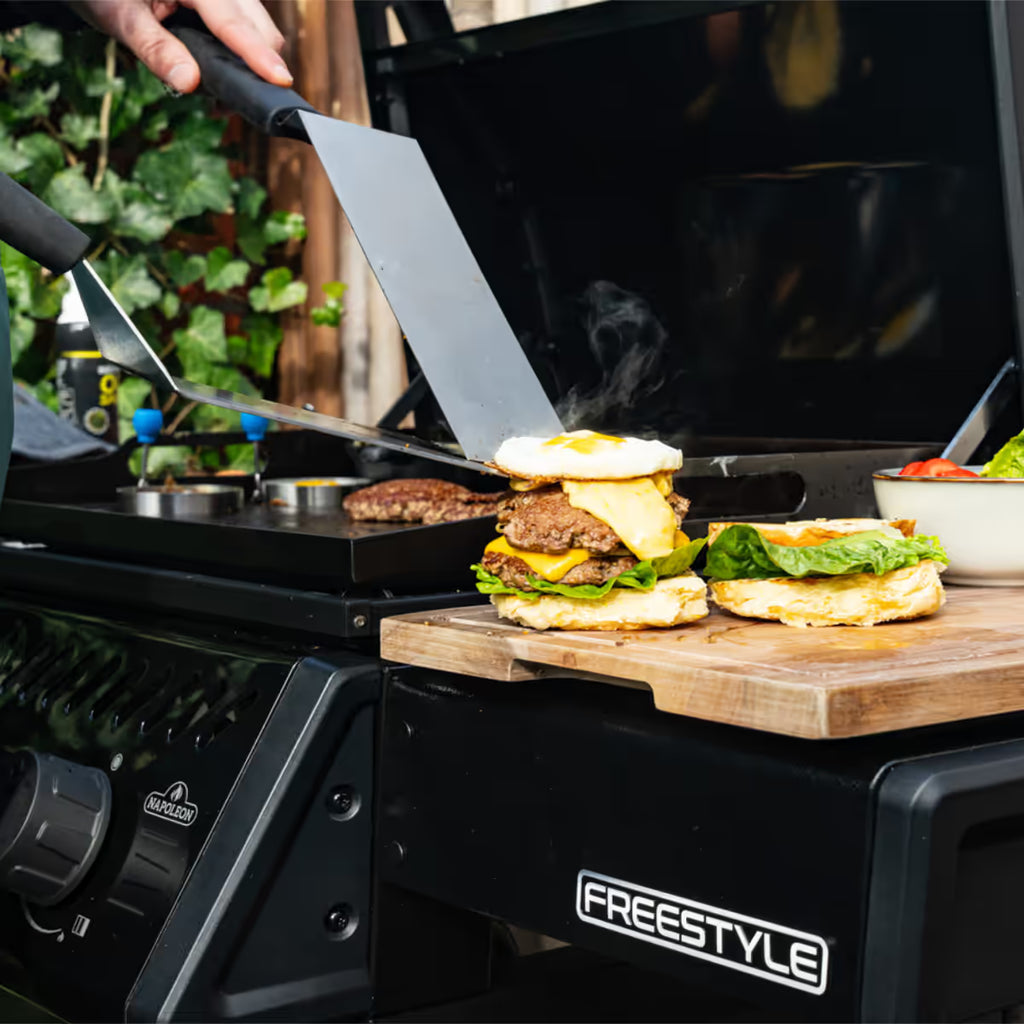 Primer plano de espátulas preparando hamburguesas con queso y huevo sobre la plancha del asador Napoleon Freestyle 24 - Ferretería La Palma - 1890000