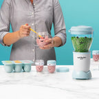 Mujer mezclando puré rosa con una cuchara amarilla en el vaso con asas de la Nutribullet Baby de 200W junto a recipientes pequeños - Ferretería La Palma - 1690012