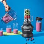 Mano virtiendo licuado desde la jarra de la Nutribullet Blender Combo 1200W hacia vasos junto a un vaso individual con frutas sobre fondo azul - Ferretería La Palma - 1690005