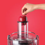 Mano introduciendo una manzana roja entera en el tubo de entrada ancho de la Nutribullet Juicer 800W sobre un fondo rojo - Ferretería La Palma - 1690019