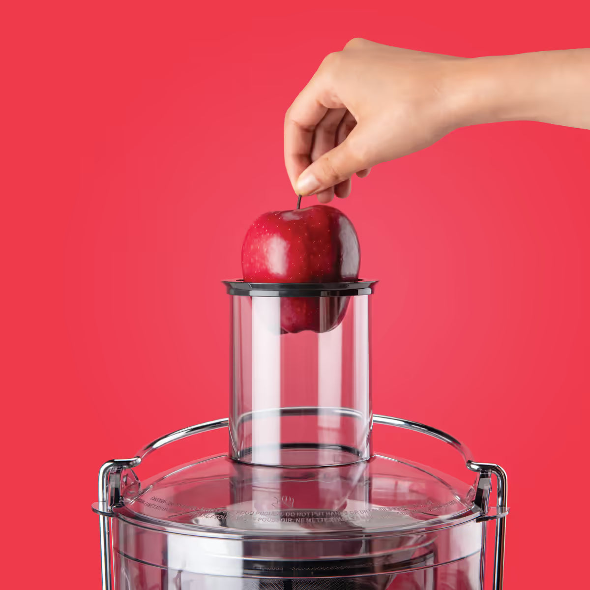 Mano introduciendo una manzana roja entera en el tubo de entrada ancho de la Nutribullet Juicer 800W sobre un fondo rojo - Ferretería La Palma - 1690019