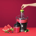 Mano introduciendo una manzana verde en el extractor Nutribullet Juicer 800W con jugo preparado al lado de frutas frescas sobre fondo rojo - Ferretería La Palma - 1690019