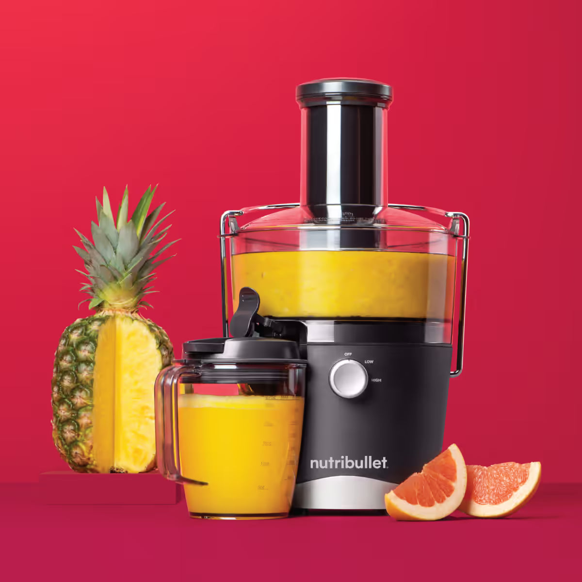 Extractor Nutribullet Juicer 800W con jugo de piña en la jarra junto a una piña cortada y rodajas de toronja sobre fondo rojo - Ferretería La Palma - 1690019