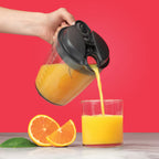 Mano virtiendo jugo de naranja desde la jarra de la Nutribullet Juicer 800W hacia un vaso junto a rodajas de fruta sobre fondo rojo - Ferretería La Palma - 1690019
