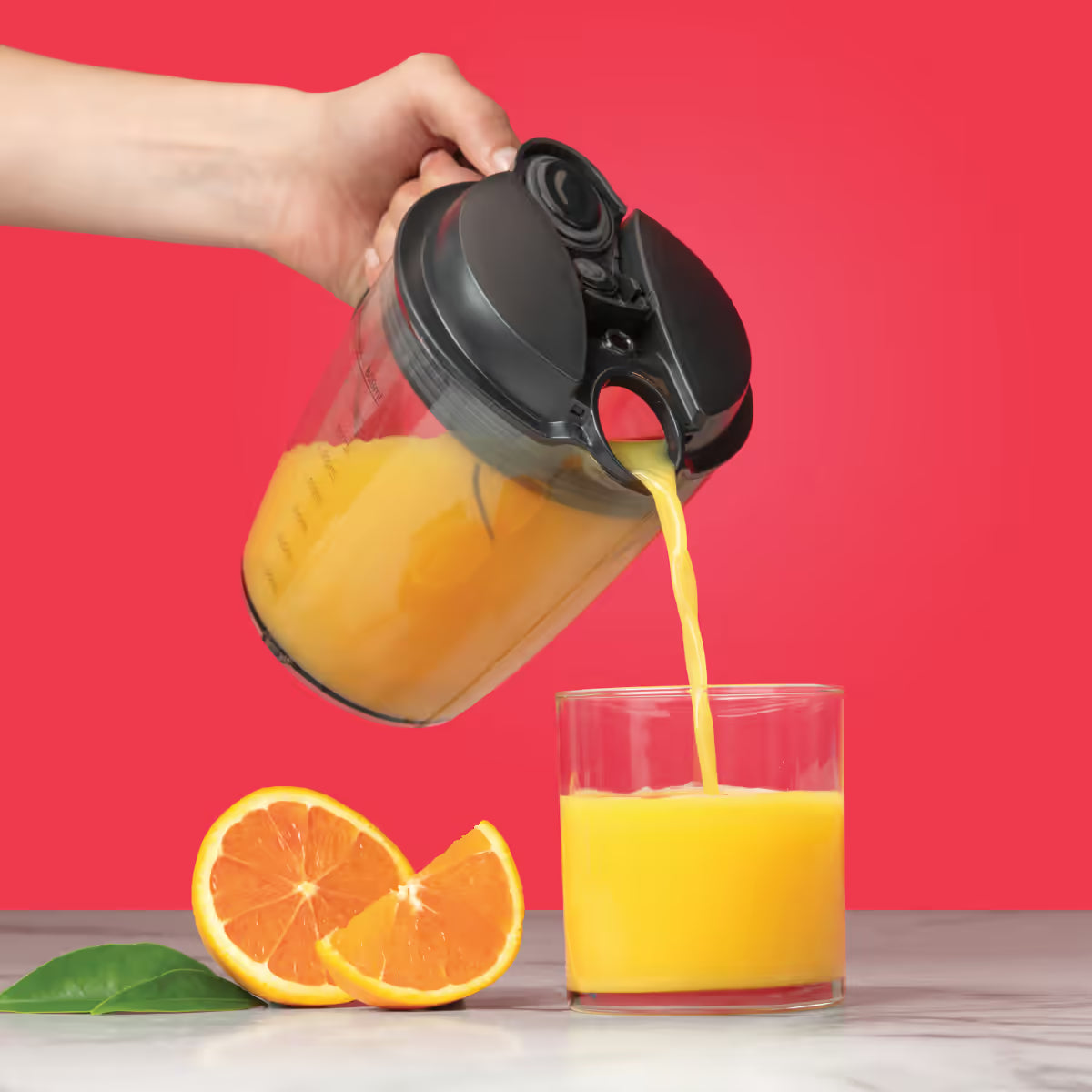Mano virtiendo jugo de naranja desde la jarra de la Nutribullet Juicer 800W hacia un vaso junto a rodajas de fruta sobre fondo rojo - Ferretería La Palma - 1690019