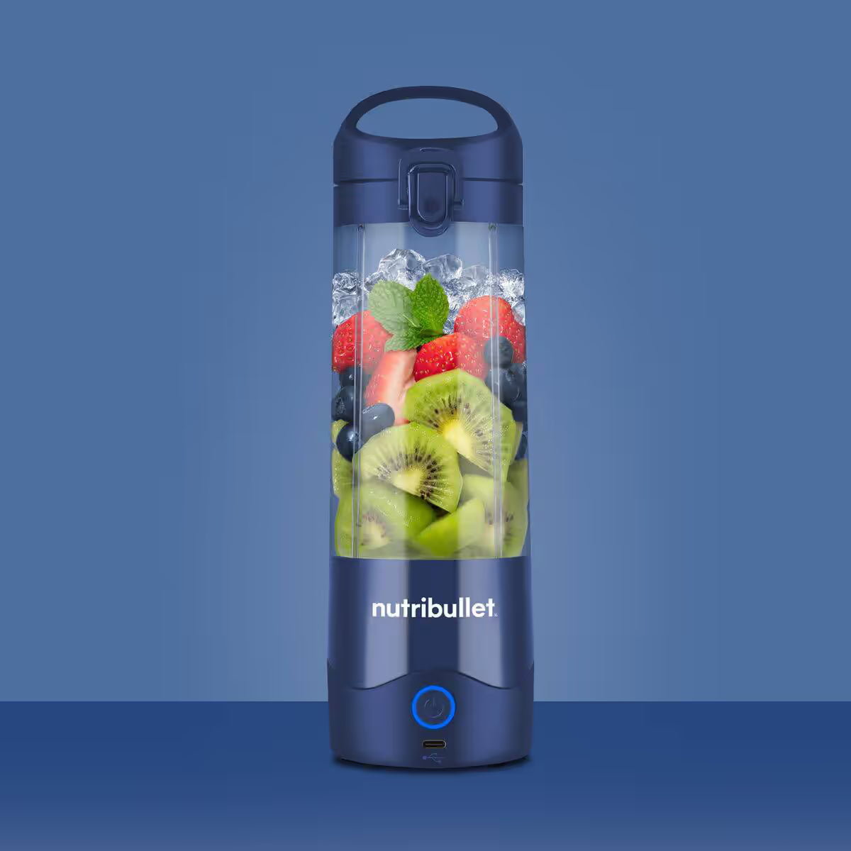 Vista frontal de la licuadora portátil Nutribullet Portable azul de 16oz con frutas e hielo sobre fondo azul - Ferretería La Palma - 1690100