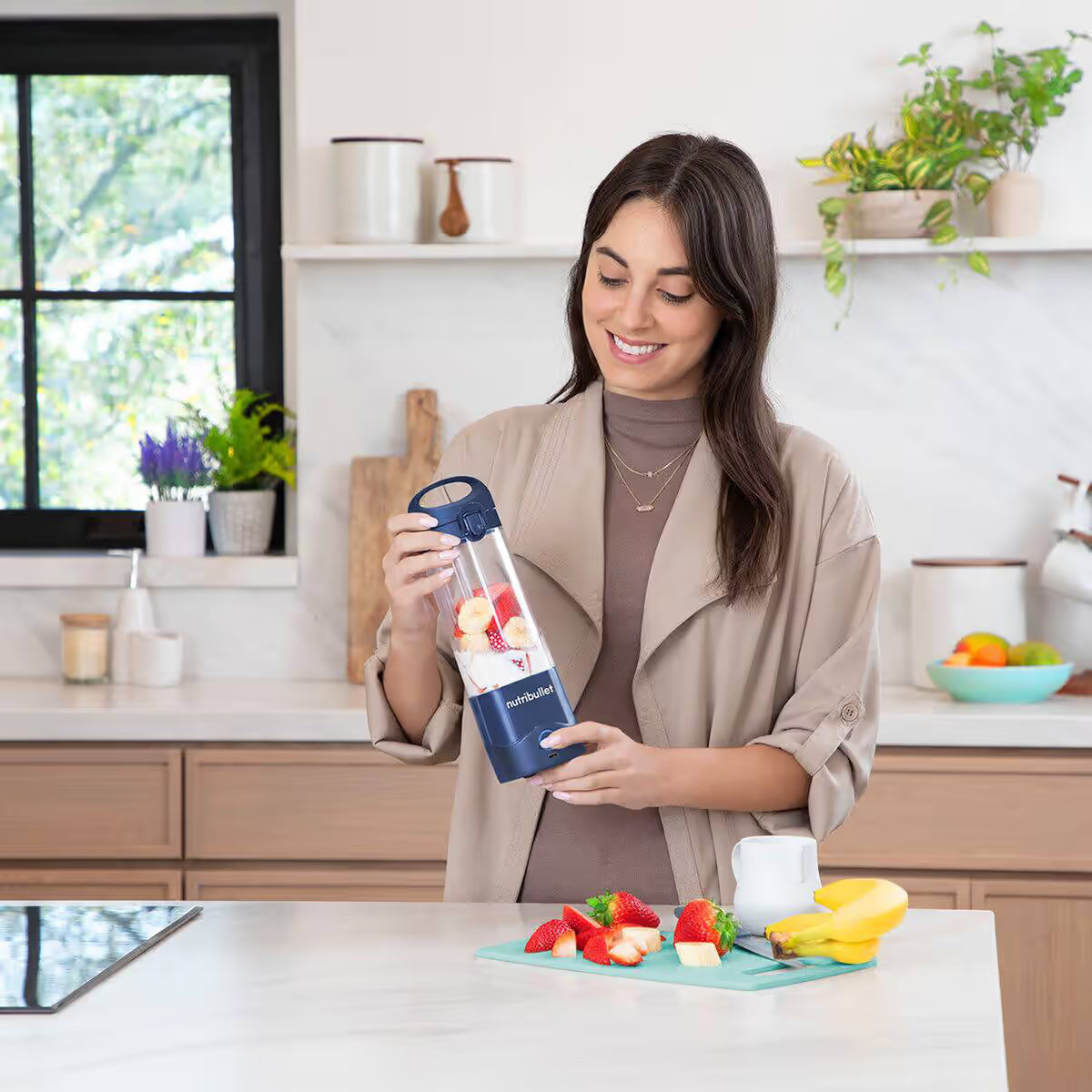 Mujer sonriendo en una cocina mientras sostiene la licuadora portátil Nutribullet Portable azul de 16oz con fresas y plátanos - Ferretería La Palma - 1690100
