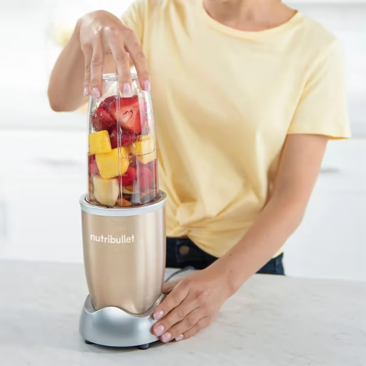 Mujer utilizando la licuadora personal Nutribullet Pro 900W en color champagne para procesar un batido de fresas y piña en una encimera de cocina - Ferretería La Palma - 1690013