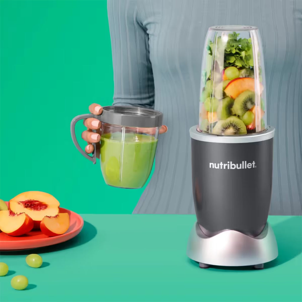 Vista frontal de la licuadora personal Nutribullet Original de 600W con kiwi y vegetales en el vaso principal, mientras una persona sostiene el vaso con anillo con asa conteniendo un licuado verde sobre un fondo turquesa - Almacén La Palma - 1690000