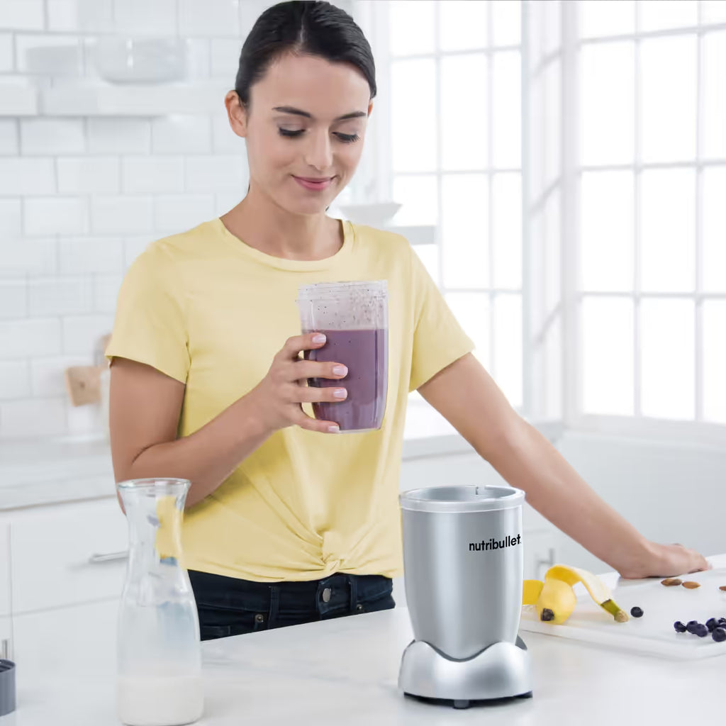 Mujer sosteniendo un vaso con licuado de frutas junto a la base motorizada de la licuadora personal Nutribullet Original 600W plateada en una cocina - Ferretería La Palma - 1690080