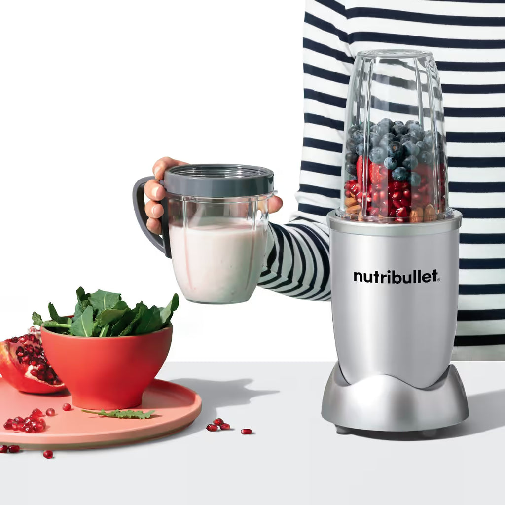 Persona sosteniendo un vaso con batido preparado junto a la licuadora personal Nutribullet Original 600W plateada con frutas sobre fondo blanco - Ferretería La Palma - 1690080