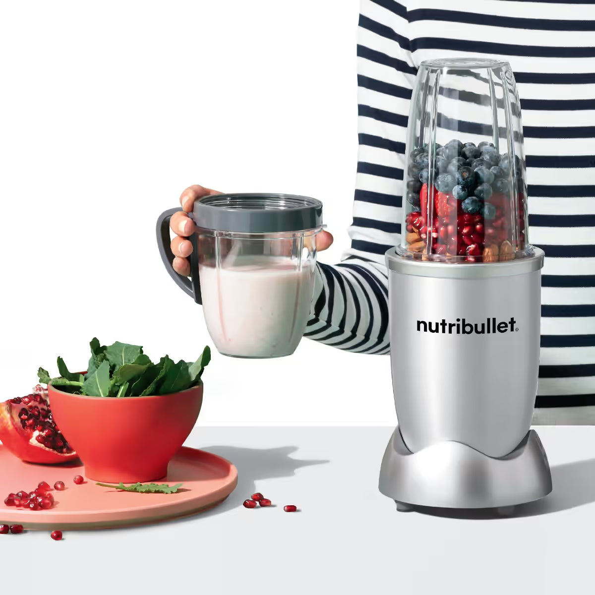 Persona sosteniendo un vaso con batido preparado junto a la licuadora personal Nutribullet Original 600W plateada con frutas sobre fondo blanco - Ferretería La Palma - 1690080