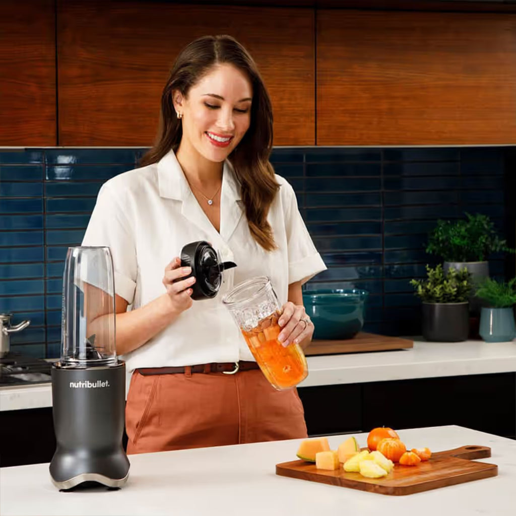 Mujer sonriendo en una cocina mientras coloca la tapa portátil al vaso con batido de frutas de la licuadora Nutribullet Ultra 1200W - Ferretería La Palma - 1690110