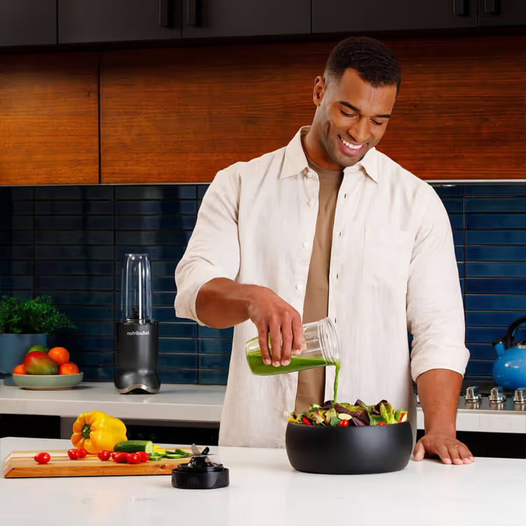 Hombre sonriendo en una cocina mientras vierte aderezo verde preparado con la licuadora Nutribullet Ultra 1200W sobre una ensalada - Ferretería La Palma - 1690110