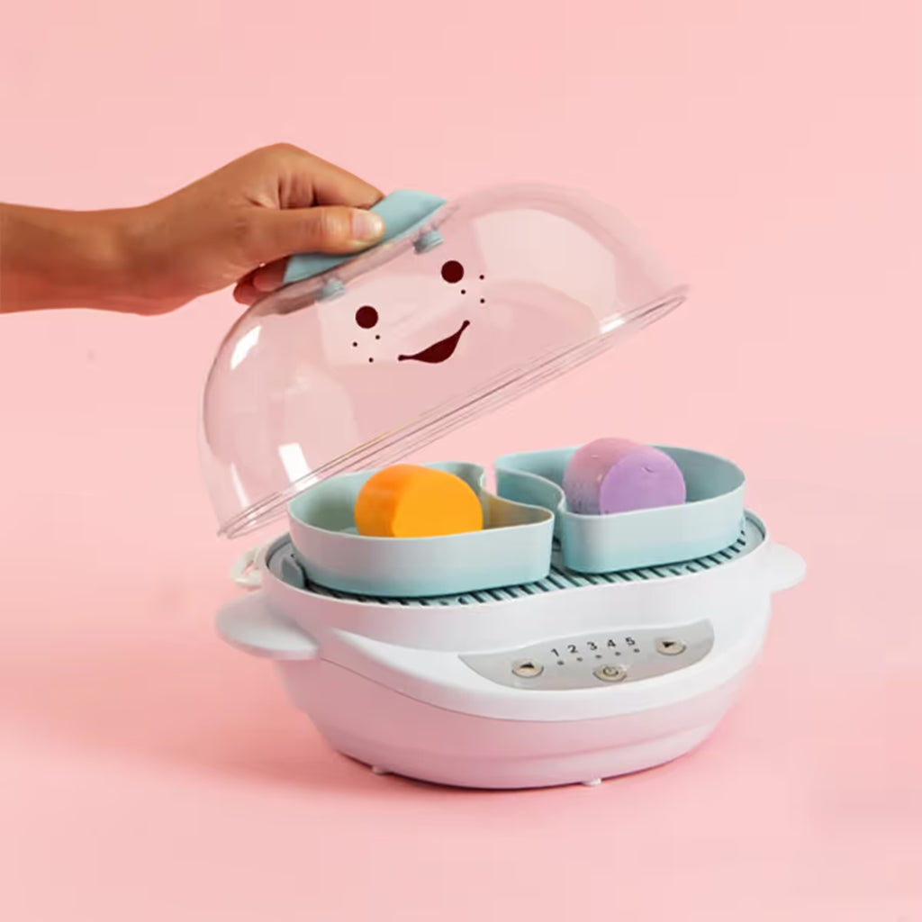 Mano levantando la tapa transparente de la vaporera Nutribullet Turbo Baby 900W para mostrar dos recipientes con alimentos sobre un fondo rosa - Ferretería La Palma - 1690014