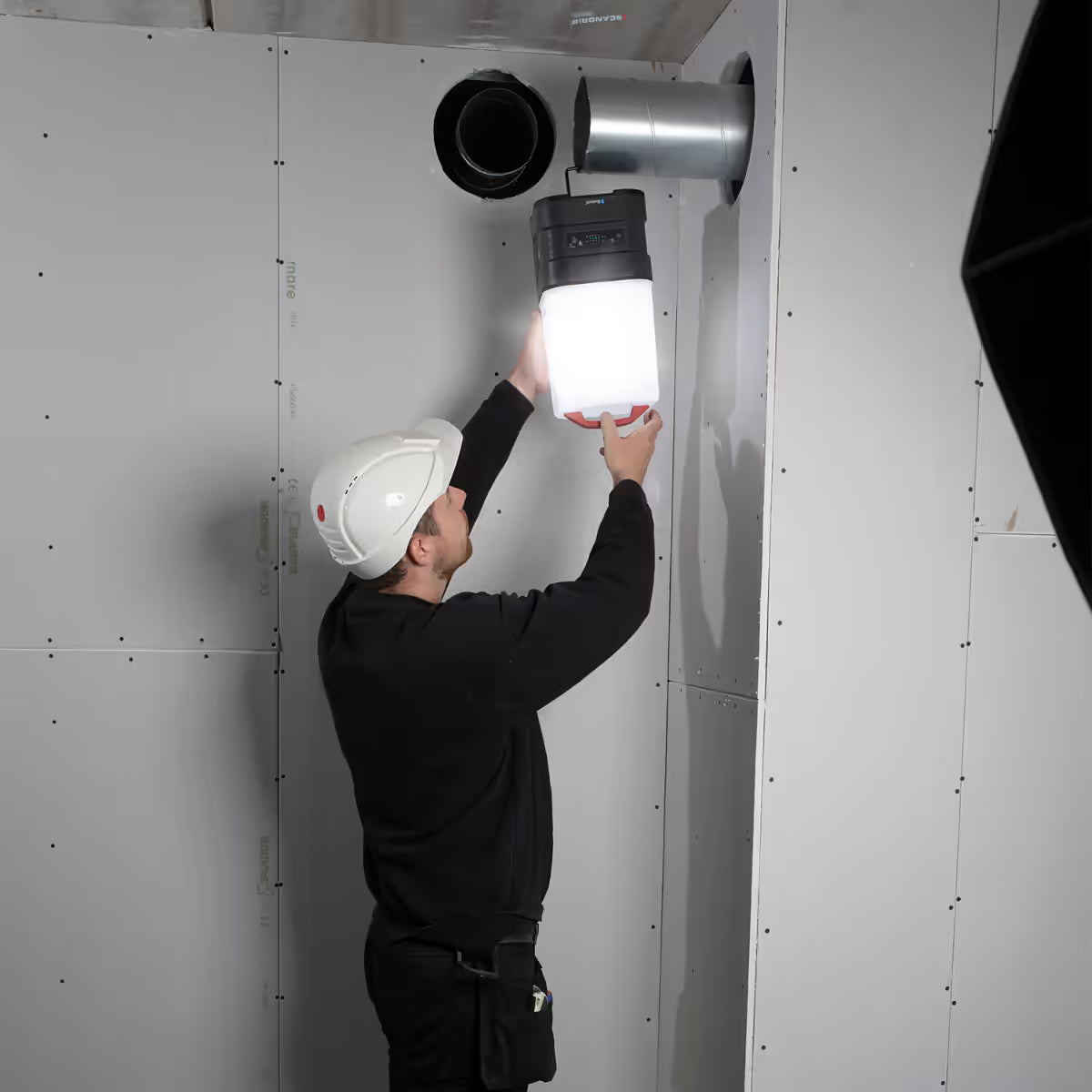 Operario con casco de seguridad colgando el reflector industrial LED Scangrip AREA 10 CONNECT de un ducto metálico para iluminar una obra - Ferretería La Palma - 1870011