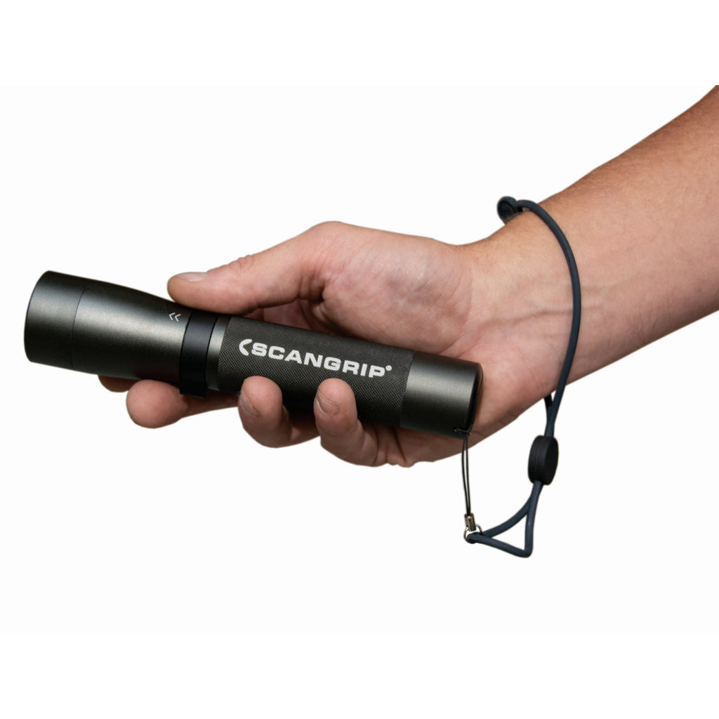 Mano sosteniendo horizontalmente la linterna LED Scangrip FLASH 600 R mostrando el agarre y la correa de muñeca sobre fondo blanco - Ferretería La Palma - 1870006