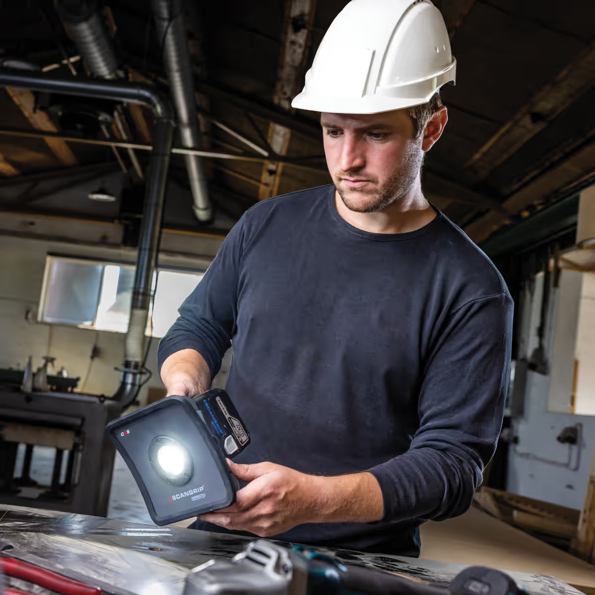 Operario con casco de seguridad utilizando el reflector LED compacto industrial Scangrip NOVA 2 CONNECT encendido para iluminar su mesa de trabajo en un taller - Ferretería La Palma - 1870008