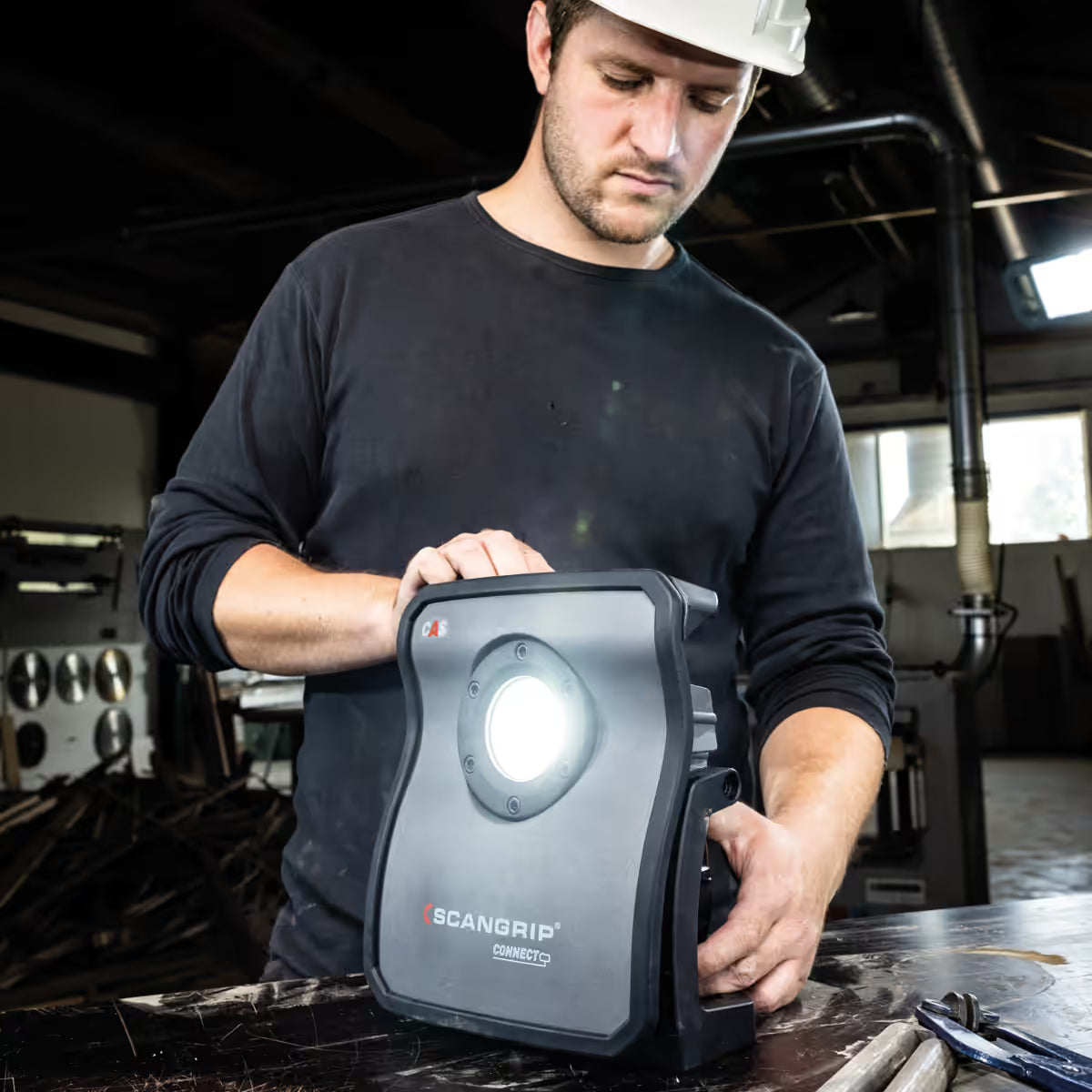 Operario con casco de seguridad encendiendo y ajustando la posición del reflector LED Scangrip NOVA 4 CONNECT sobre su mesa de trabajo en un taller - Ferretería La Palma - 1870009