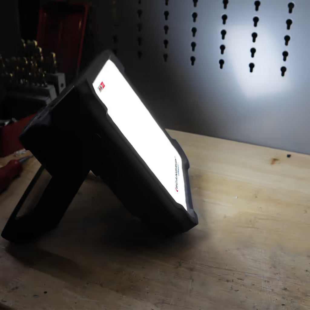 Reflector LED compacto Scangrip VEGA 2 CONNECT encendido sobre una mesa de trabajo proyectando luz difusa hacia un panel organizador metálico - Ferretería La Palma - 1870012