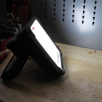 Reflector LED compacto Scangrip VEGA 2 CONNECT encendido sobre una mesa de trabajo proyectando luz difusa hacia un panel organizador metálico - Ferretería La Palma - 1870012