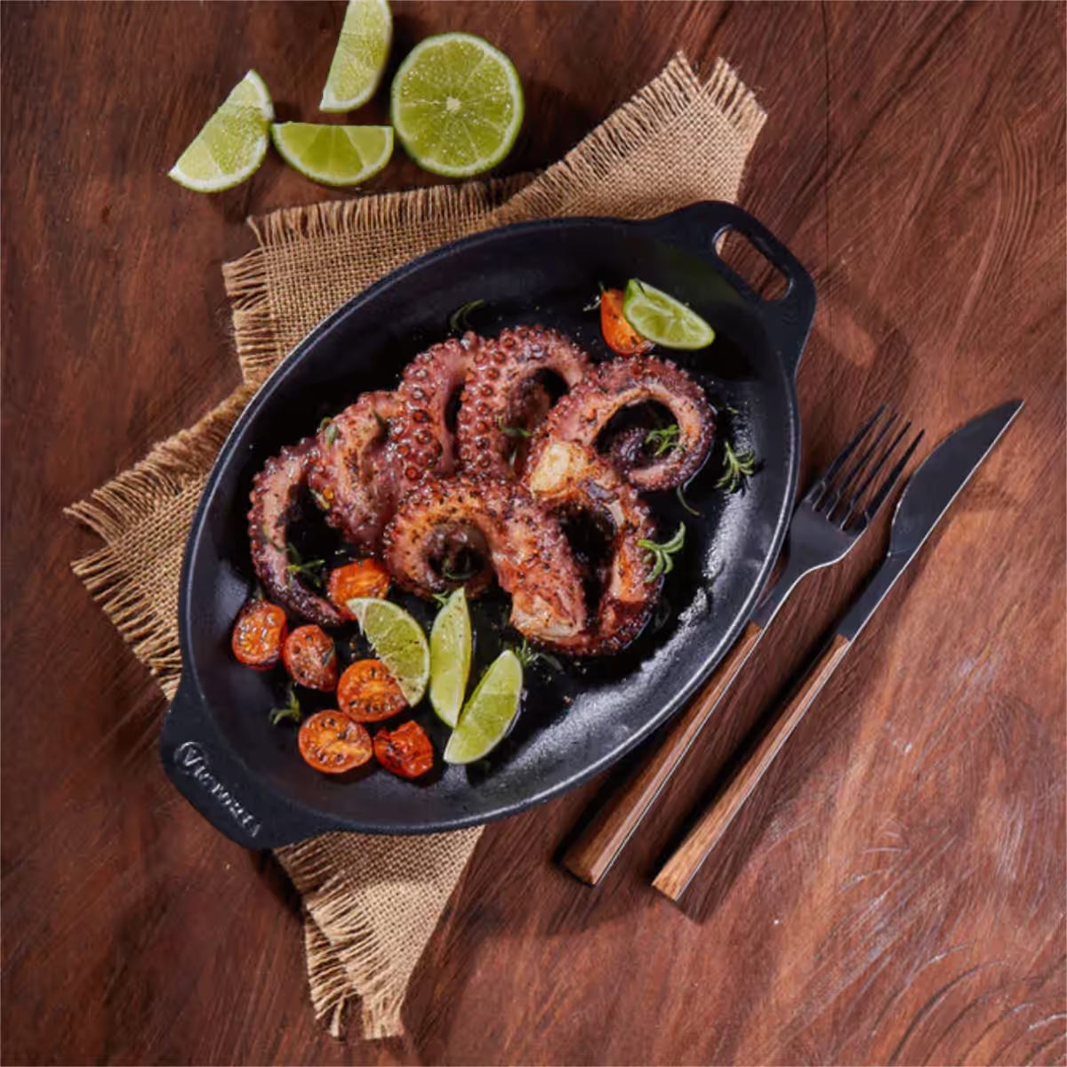 Vista cenital de la bandeja ovalada Victoria de 29x19 cm en hierro fundido esmaltado con pulpo asado, tomates cherry y limones sobre una mesa de madera con cubiertos - Ferretería La Palma - 1050041