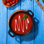 Vista cenital de la cazuela redonda Victoria de 14 cm en hierro fundido esmaltado con sopa de tomate decorada con crema servida sobre una base de madera en una mesa azul - Ferretería La Palma - 1050042