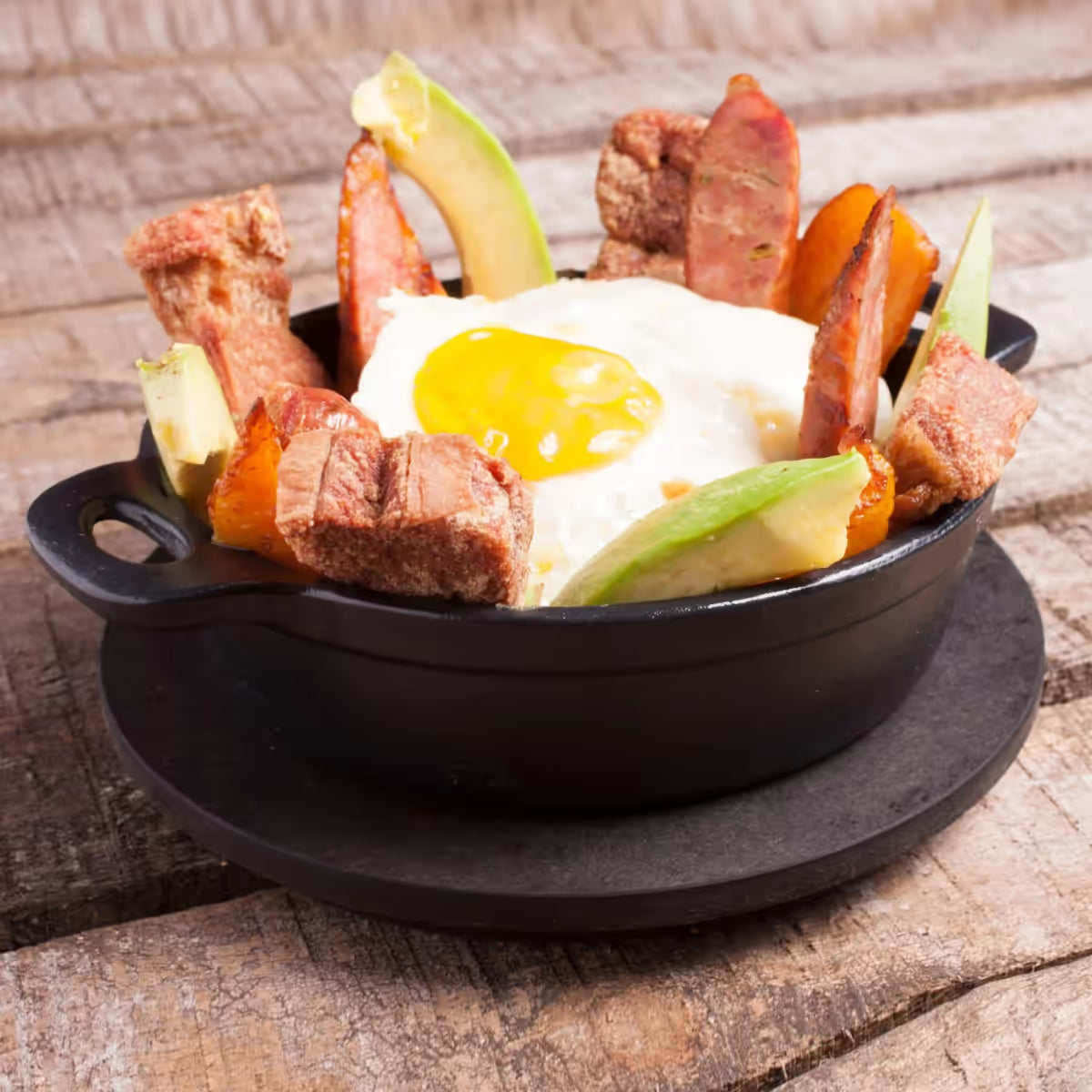 Vista en perspectiva de la cazuela redonda Victoria de 16 cm en hierro fundido esmaltado servida con chicharrón, huevo frito, aguacate y chorizo sobre una base negra en una mesa de madera - Ferretería La Palma - 1050043