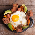 Vista cenital de la cazuela redonda Victoria de 16 cm en hierro fundido esmaltado servida con huevo frito, chicharrón, aguacate, chorizo y plátano maduro sobre una base negra en una mesa de madera - Ferretería La Palma - 1050043