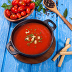 Vista en perspectiva de la cazuela redonda Victoria de 16 cm en hierro fundido esmaltado con sopa de tomate decorada con hierbas y crutones servida sobre una base de madera en una mesa azul - Ferretería La Palma - 1050043