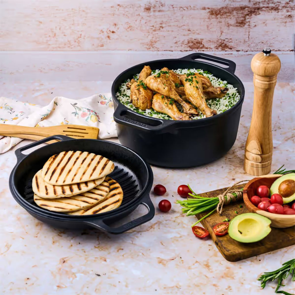 Vista superior en perspectiva del Combo Cooker 2 en 1 Victoria de 5.7 litros en hierro fundido esmaltado con pollo asado sobre arroz en la olla y panes asados en la tapa tipo grill, acompañados de vegetales frescos sobre una superficie clara - Ferretería La Palma - 1050079