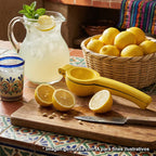 Exprimidor de limones manual Victoria de aluminio fundido color amarillo sobre una tabla de madera con limones cortados, junto a una jarra de limonada fresca y una canasta de limones en un ambiente de cocina - Ferretería La Palma - 1050001