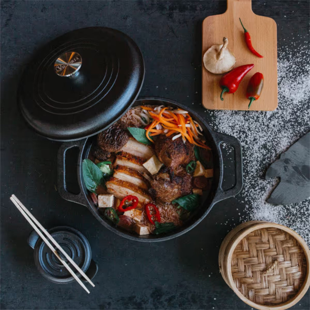 Vista superior en perspectiva del horno holandés Victoria de 3.8 litros en hierro fundido curado con una sopa de fideos, panceta de cerdo y vegetales, acompañada de una tabla con chiles y una vaporera de bambú sobre una superficie oscura - Ferretería La Palma - 1050045