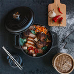 Vista superior en perspectiva del horno holandés Victoria de 3.8 litros en hierro fundido esmaltado con una sopa de fideos, panceta de cerdo y vegetales, acompañada de una tabla con chiles y una vaporera de bambú sobre una superficie oscura - Ferretería La Palma - 1050044