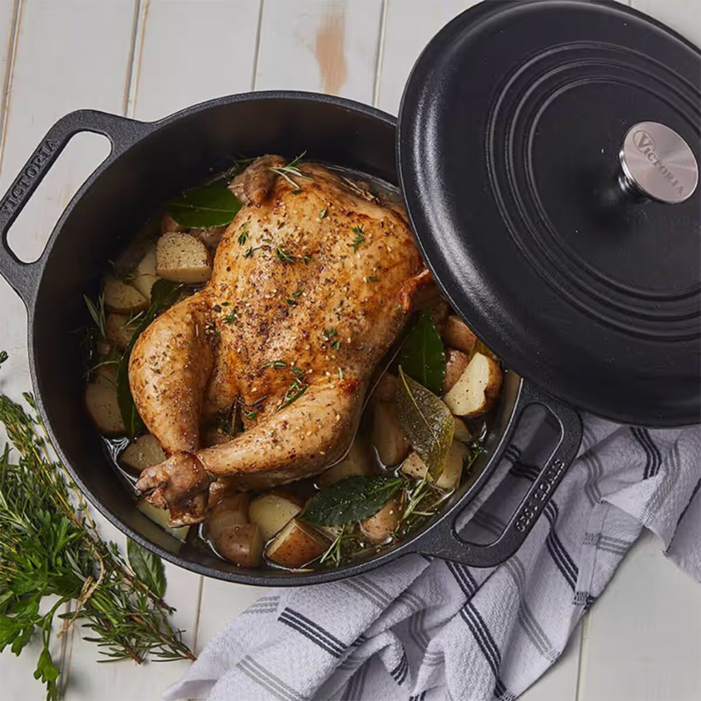 Vista superior en perspectiva del horno holandés Victoria de 6.6 litros en hierro fundido esmaltado con un pollo entero asado, papas y hierbas aromáticas sobre una mesa de madera blanca - Ferretería La Palma - 1050085