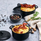 Vista en perspectiva de dos mini ollas cocotte redondas Victoria de 0.3 L en hierro fundido esmaltado servidas con un gratinado y tocino crujiente, acompañadas de pimienta, nuez moscada y romero sobre una superficie gris - Ferretería La Palma - 1050059