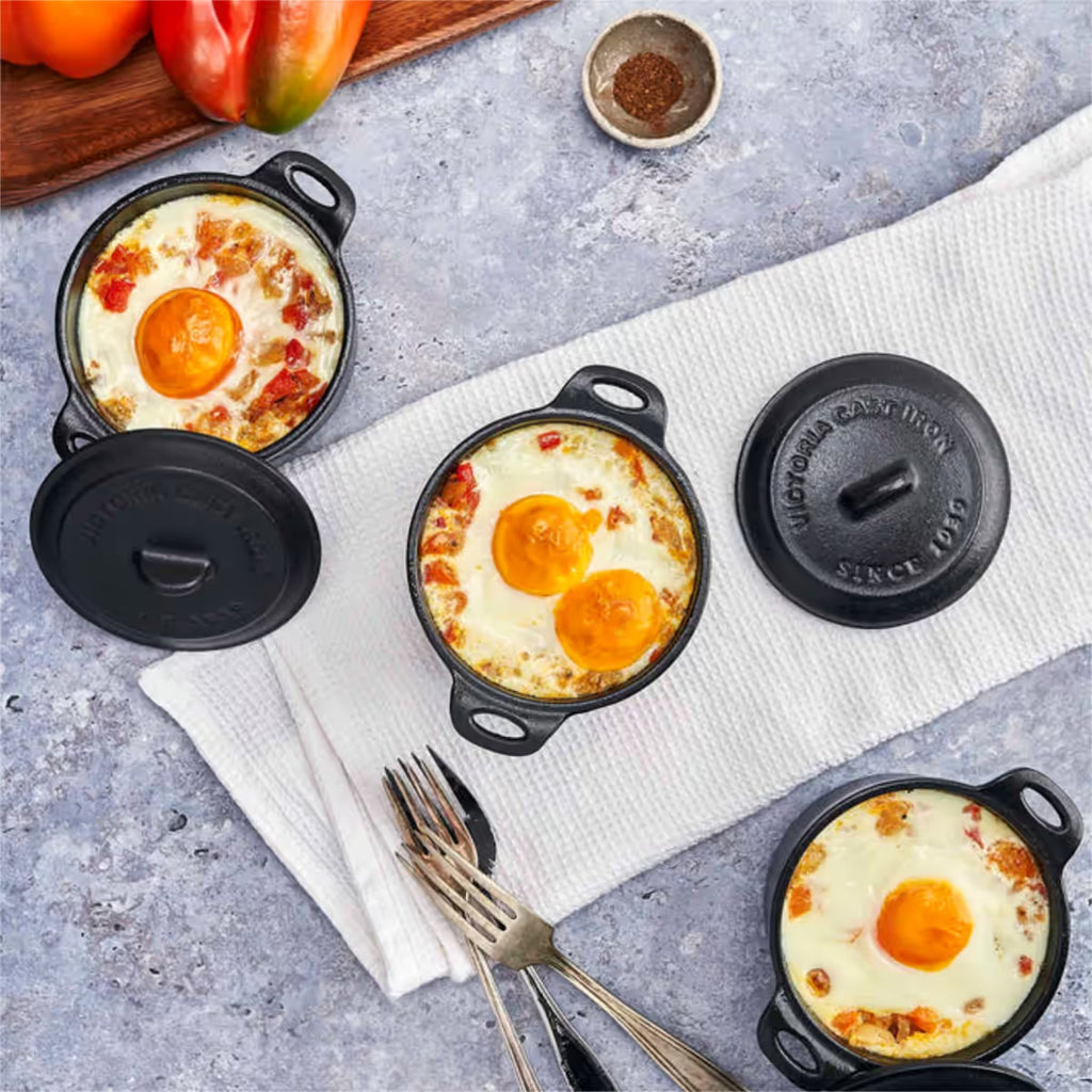 Vista superior de tres mini ollas cocotte redondas Victoria de 0.3 L en hierro fundido esmaltado servidas con huevos al horno y vegetales, acompañadas de cubiertos y pimientos sobre una superficie gris - Ferretería La Palma - 1050059