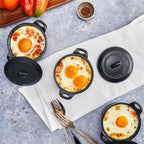 Vista superior de tres mini ollas cocotte redondas Victoria de 0.3 L en hierro fundido esmaltado servidas con huevos al horno y vegetales, acompañadas de cubiertos y pimientos sobre una superficie gris - Ferretería La Palma - 1050059