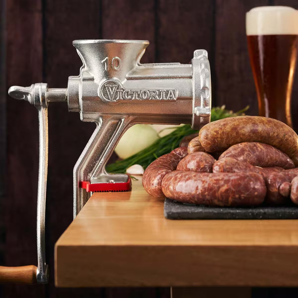Molino de carne manual Victoria número 10 de hierro fundido instalado en mesa de madera, junto a embutidos artesanales y vegetales, ideal para preparar chorizos y molienda doméstica - Ferretería La Palma - 1440002