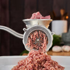 Primer plano frontal del molino de carne manual Victoria número 12 en uso, mostrando la salida de carne molida a través del disco de acero hacia una bandeja blanca, con carne fresca en la tolva superior - Ferretería La Palma - 1440003