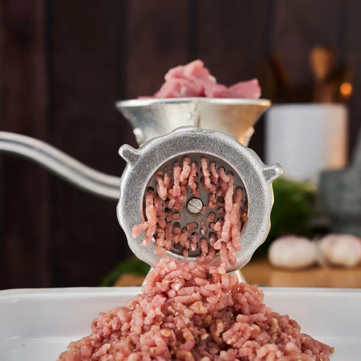 Primer plano frontal del molino de carne manual Victoria número 12 en uso, mostrando la salida de carne molida a través del disco de acero hacia una bandeja blanca, con carne fresca en la tolva superior - Ferretería La Palma - 1440003