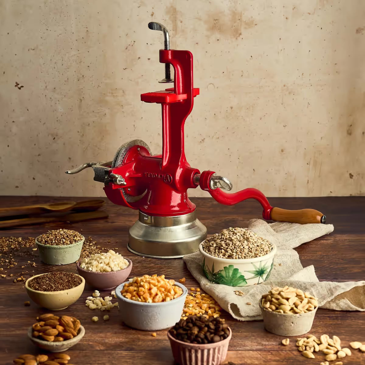 Molino de granos manual Victoria rojo de hierro fundido rodeado de diversos ingredientes como granos de café, maní, almendras y maíz, destacando su versatilidad para diferentes tipos de molienda - Ferretería La Palma - 1040046