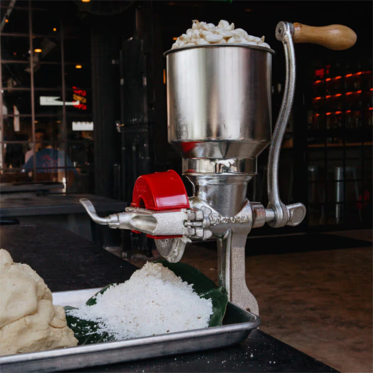 Molino de granos manual Victoria con tolva alta de 1.5 litros fabricado en hierro fundido estañado, procesando masa de maíz sobre una bandeja metálica en una cocina, destacando su capacidad de molienda fina y eficiencia - Ferretería La Palma - 1040006