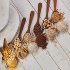 Variedad de granos, semillas y café procesados con el molino Victoria, presentados en cucharas de madera sobre fondo rústico. Muestra la versatilidad de molienda desde texturas gruesas hasta finas (maíz, maní, café, nueces). Producto 1040006 - Ferretería La Palma.