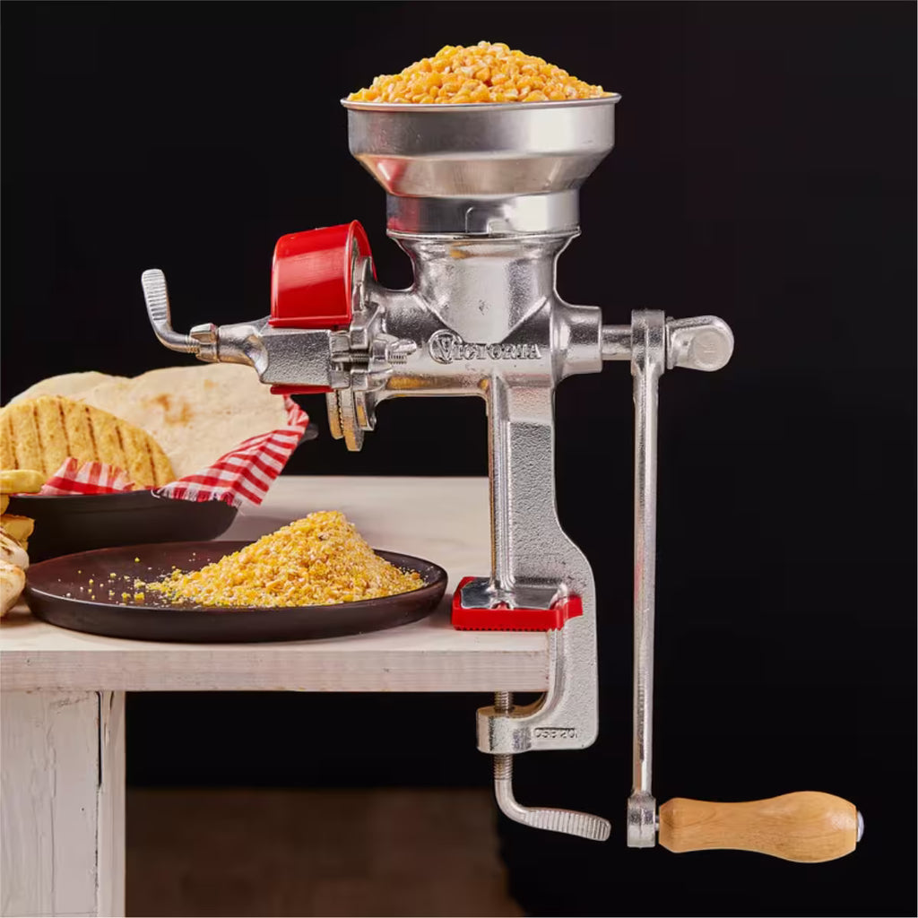 Molino de granos manual Victoria con tolva baja metálica en funcionamiento, instalado sobre una mesa blanca moliendo maíz amarillo, con arepas frescas de fondo y protector de molienda rojo - Ferretería La Palma - 1440005