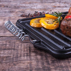 Vista de primer plano de la cara estriada tipo grill de la plancha reversible Victoria de 33x21 cm en hierro fundido esmaltado con detalle de su asa de alambre, vegetales asados y romero sobre una mesa de madera - Ferretería La Palma - 1050080