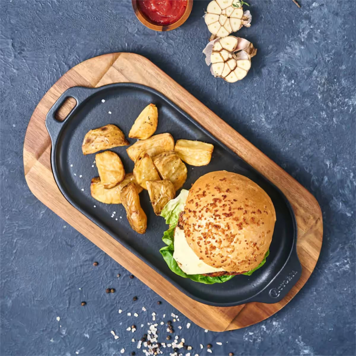Vista cenital de la plancha ovalada Victoria de 34x17 cm en hierro fundido esmaltado con una hamburguesa y papas servidas sobre una base de madera - Ferretería La Palma - 1050040