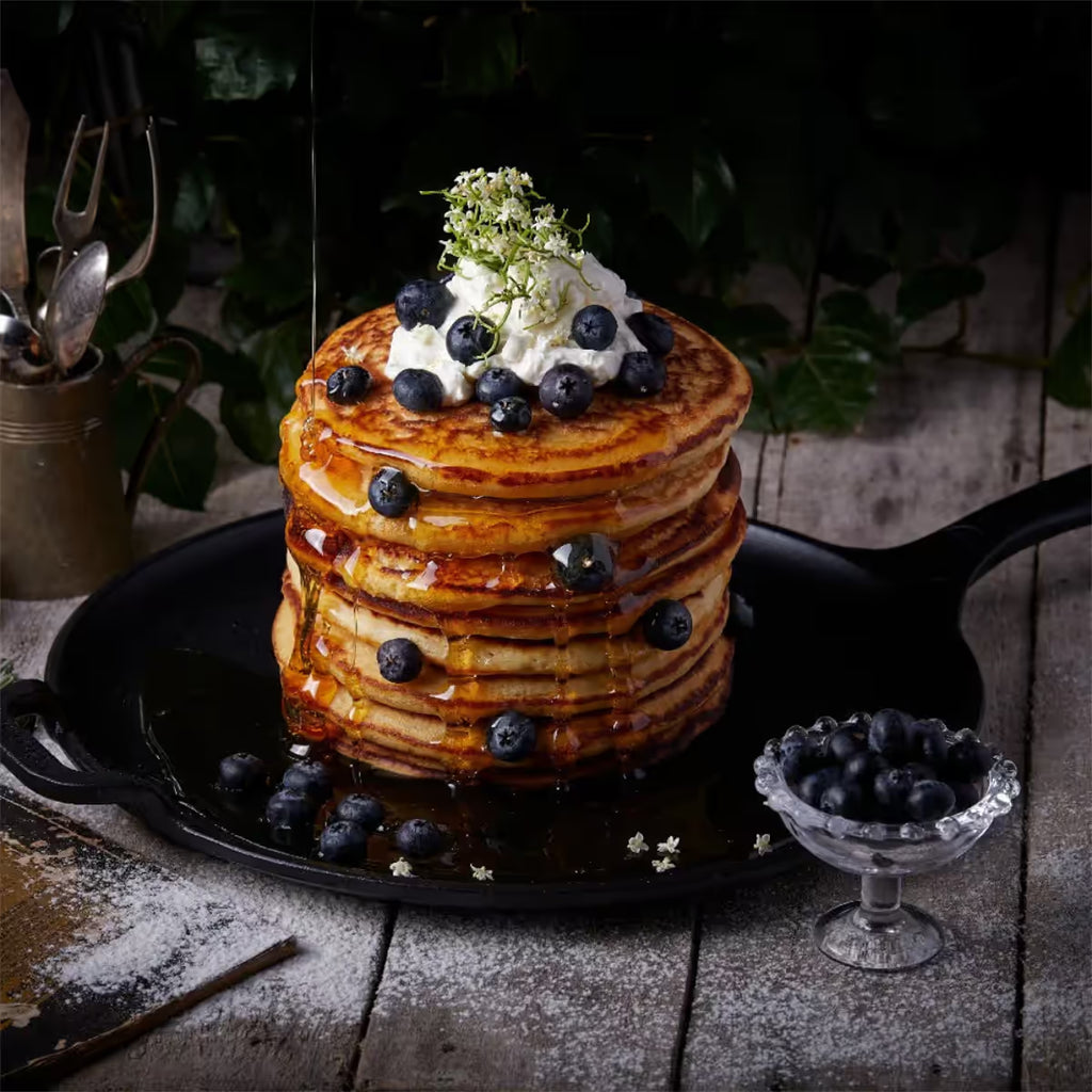 Vista en perspectiva de la plancha redonda para pizza Victoria de 30 cm en hierro fundido esmaltado con una torre de pancakes con crema, arándanos y miel sobre una mesa de madera rústica - Ferretería La Palma - 1050087