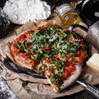 Vista en perspectiva de la plancha redonda para pizza y crepas Victoria de 25 cm en hierro fundido esmaltado con una pizza servida sobre papel pergamino, acompañada de harina, queso y aceite de oliva - Ferretería La Palma - 1050086